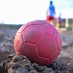 Handball liegt im Sand, im Hintergrund sind unscharf Personen zu sehen.